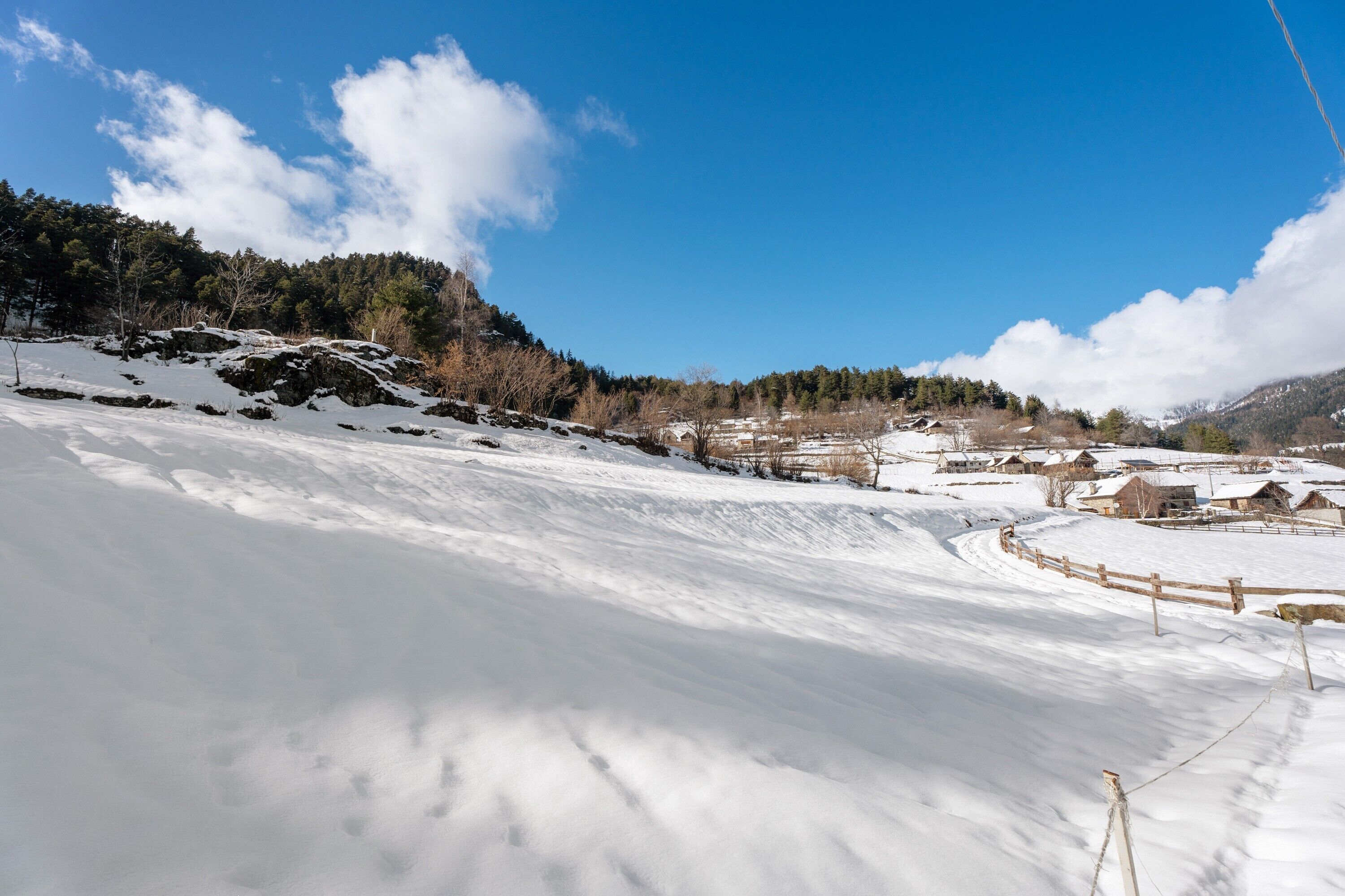 A snowy landscape with a clear sky, a wooden fence, and buildings in the distance.