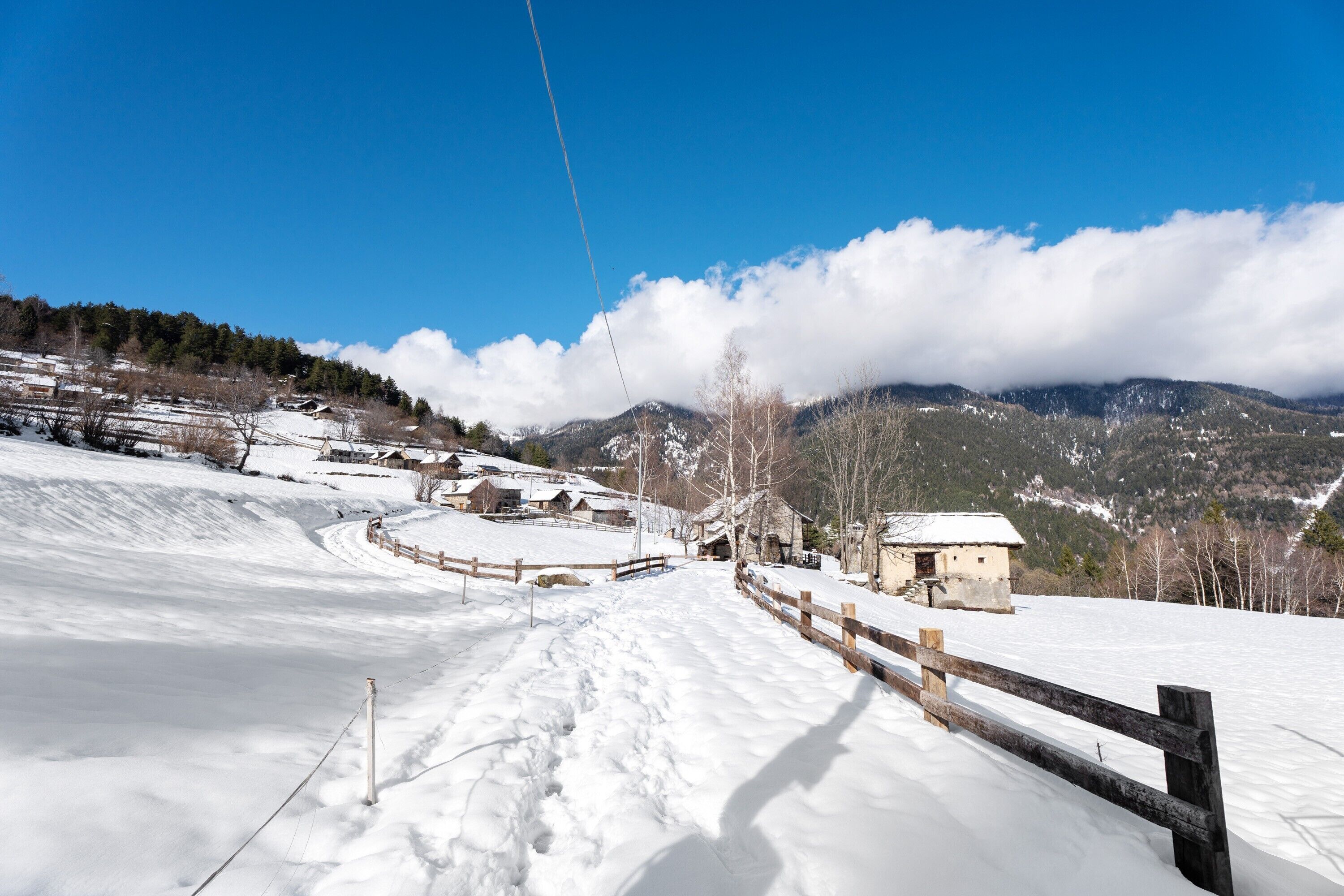A snow-covered landscape with a wooden fence, buildings, and mountains in the background.