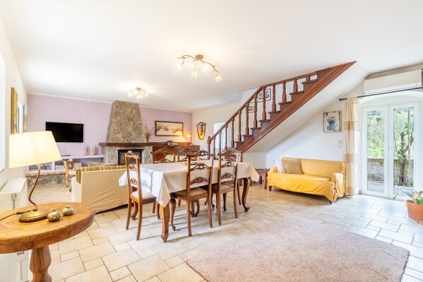 A living room with a dining table, wooden chairs, a sofa, a staircase, and a fireplace.