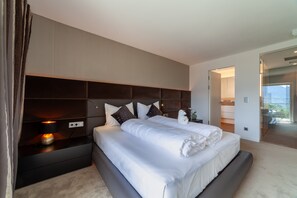 A modern bedroom with a large bed, a headboard with built-in shelves, bedside tables, and a view of the balcony.