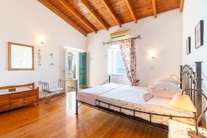 A bedroom with a wooden floor, a bed with a metal frame, a bedside lamp, a wooden bench, a mirror, and a window with curtains.