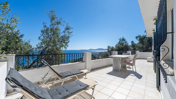 Eine Terrasse mit Liegestühlen und einem Tisch, mit Blick auf ein Gewässer und Hügel.