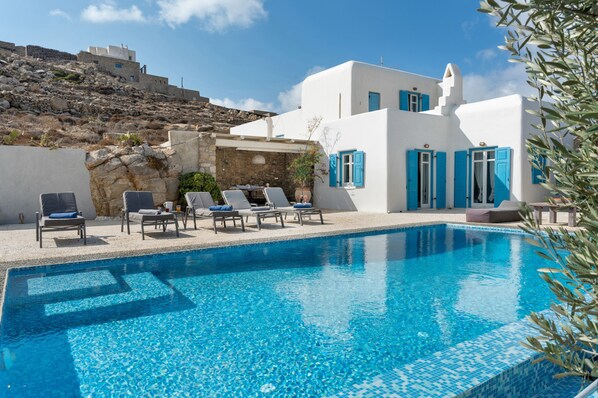Una piscina con sillas de descanso, un muro de piedra y un edificio con contraventanas azules.