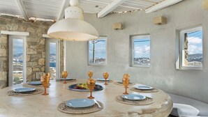 Mesa redonda para comer con platos, vasos y un frutero, frente a una pared de piedra con ventanas que ofrecen una vista.