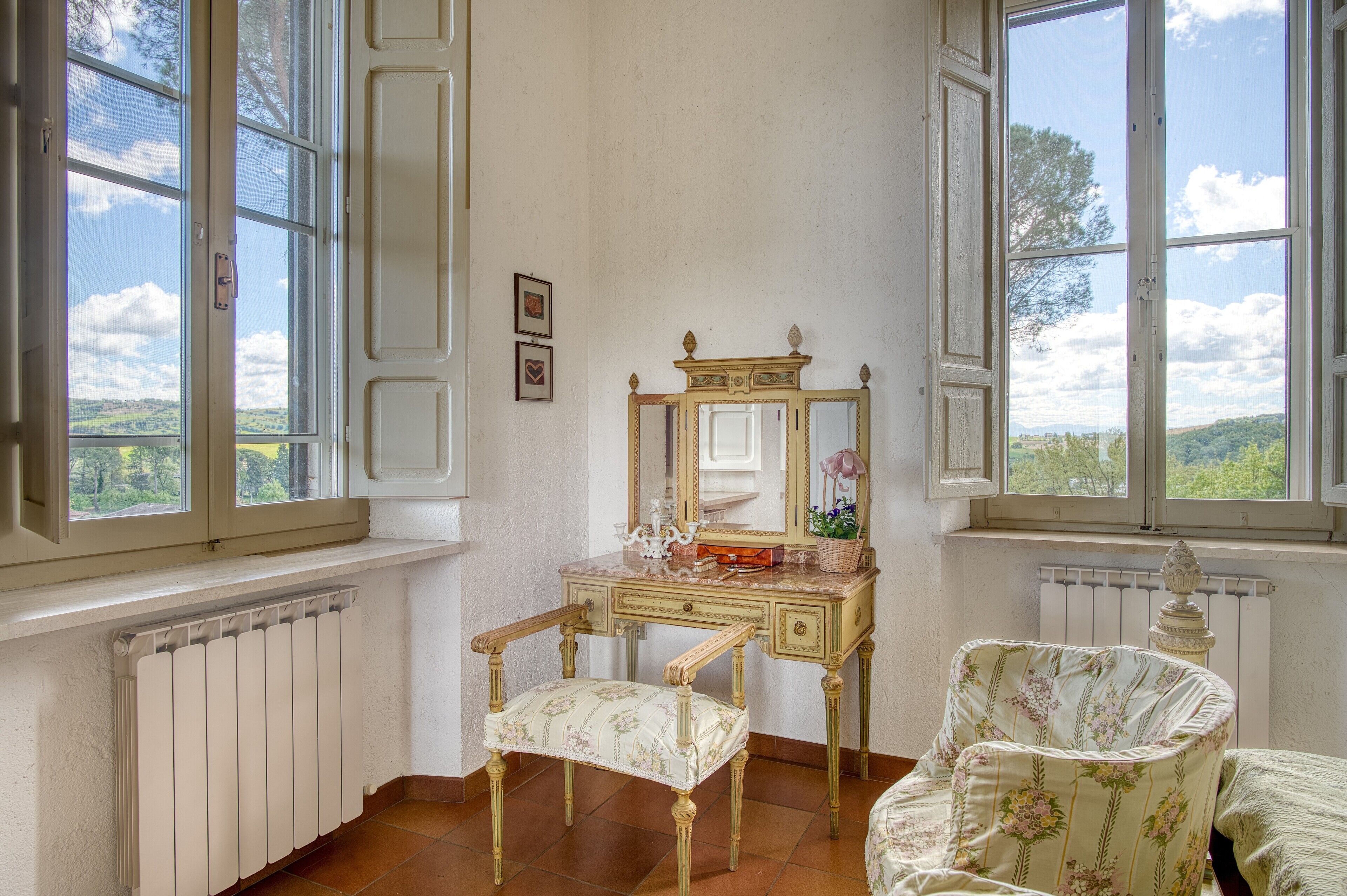 Ein Zimmer mit Kommode, Stuhl und Fenster mit Blick auf Grünflächen.
