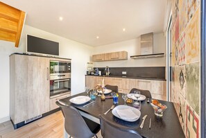 A modern kitchen with a dining table set for four, equipped with a built-in oven and microwave, and a large flat-screen TV mounted above the counter. - Maison récente à 300 m de la mer*** (Carantec)