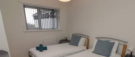 A twin bedroom with two beds, a window with blinds, a bedside table with a lamp, and a radiator.