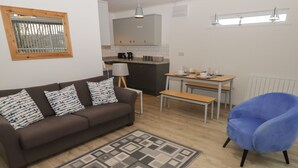 A modern living room with a brown sofa, patterned cushions, a blue armchair, a dining table with chairs, and a kitchen in the background. - 19 Coedrath Park (Saundersfoot)