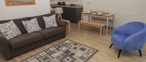 A modern living room with a brown sofa, patterned cushions, a blue armchair, a dining table with chairs, and a kitchen in the background.