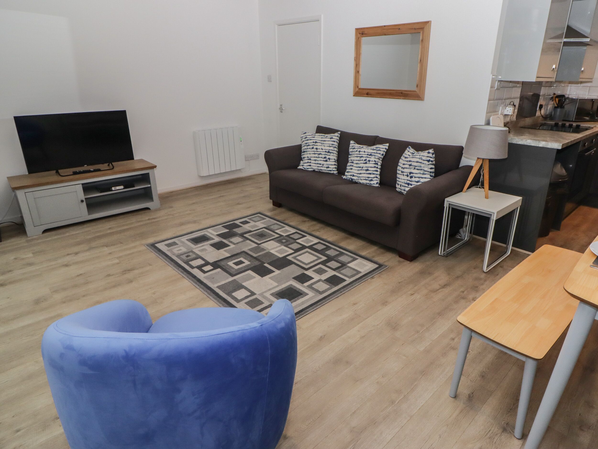A living room with a sofa, a blue armchair, a TV, a small table, and a kitchen area.