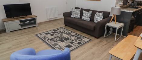 A living room with a sofa, a blue armchair, a TV, a small table, and a kitchen area.