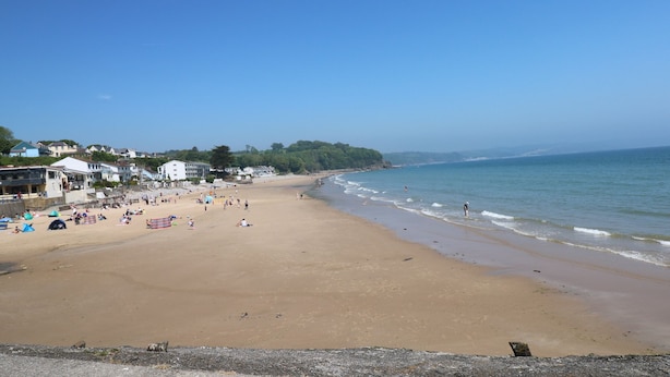 Une vaste plage de sable fin, un ciel bleu dégagé, quelques personnes et des bâtiments en arrière-plan.