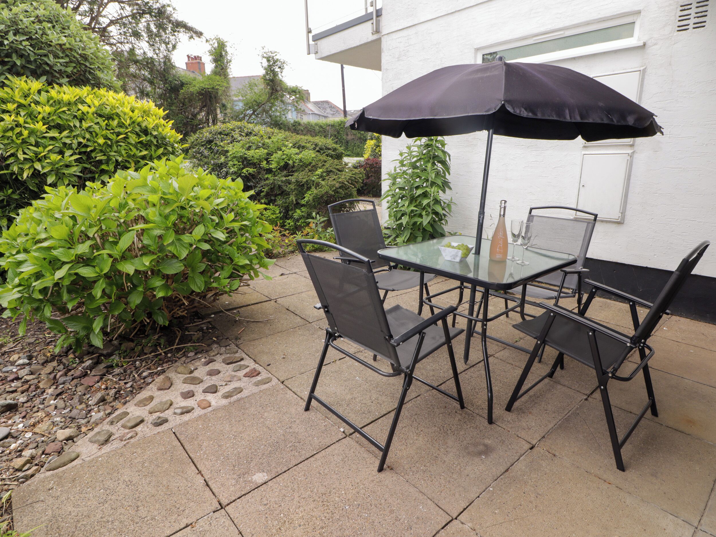 Une terrasse extérieure avec une table en verre et quatre chaises, un parasol noir et une bouteille de vin.