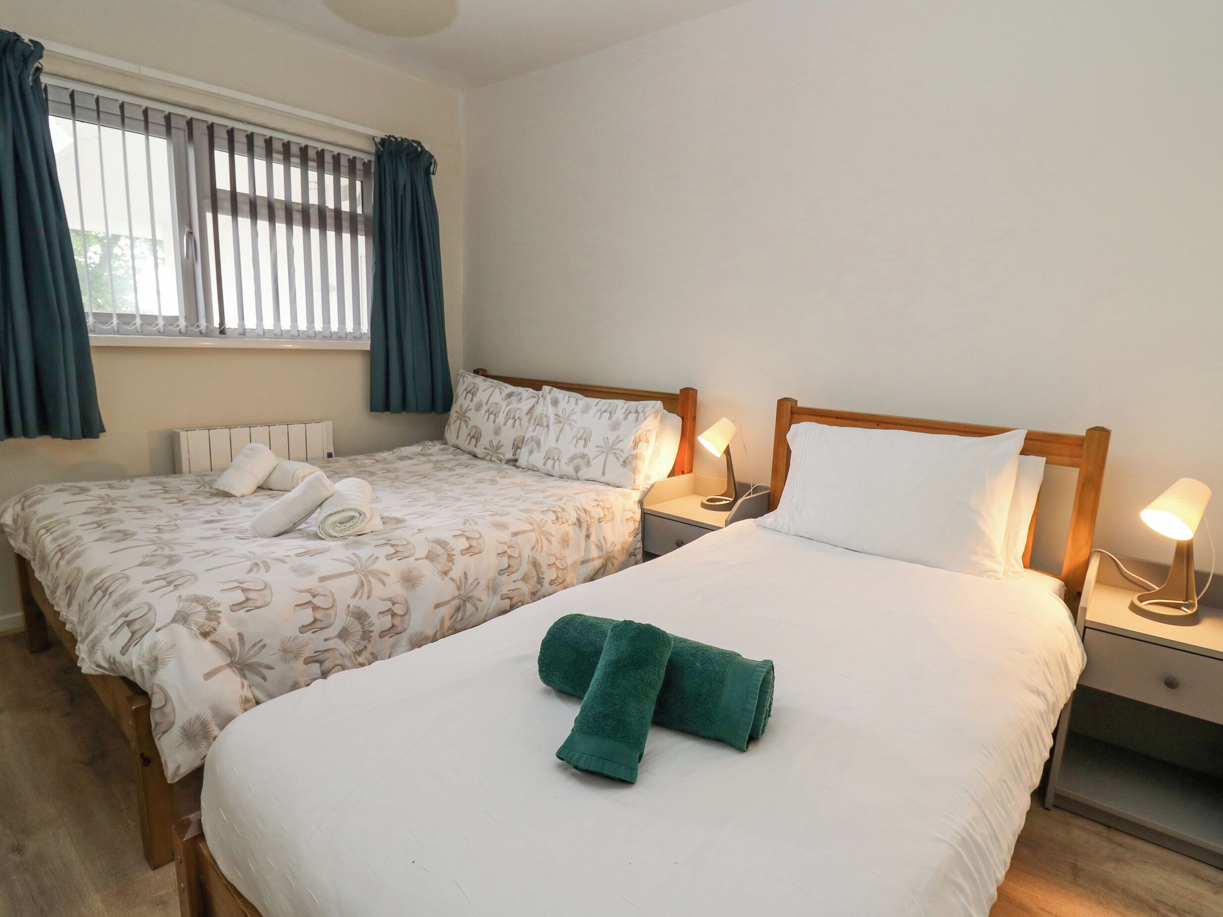 A hotel room with two beds, each with a wooden headboard, a nightstand with a lamp, and a window with curtains.