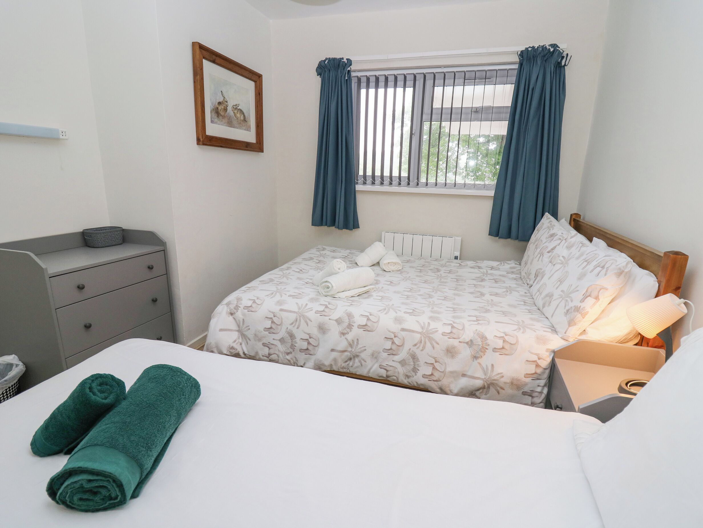 A neatly arranged bedroom with a bed, a bedside table with a lamp, a dresser, and a window with curtains.