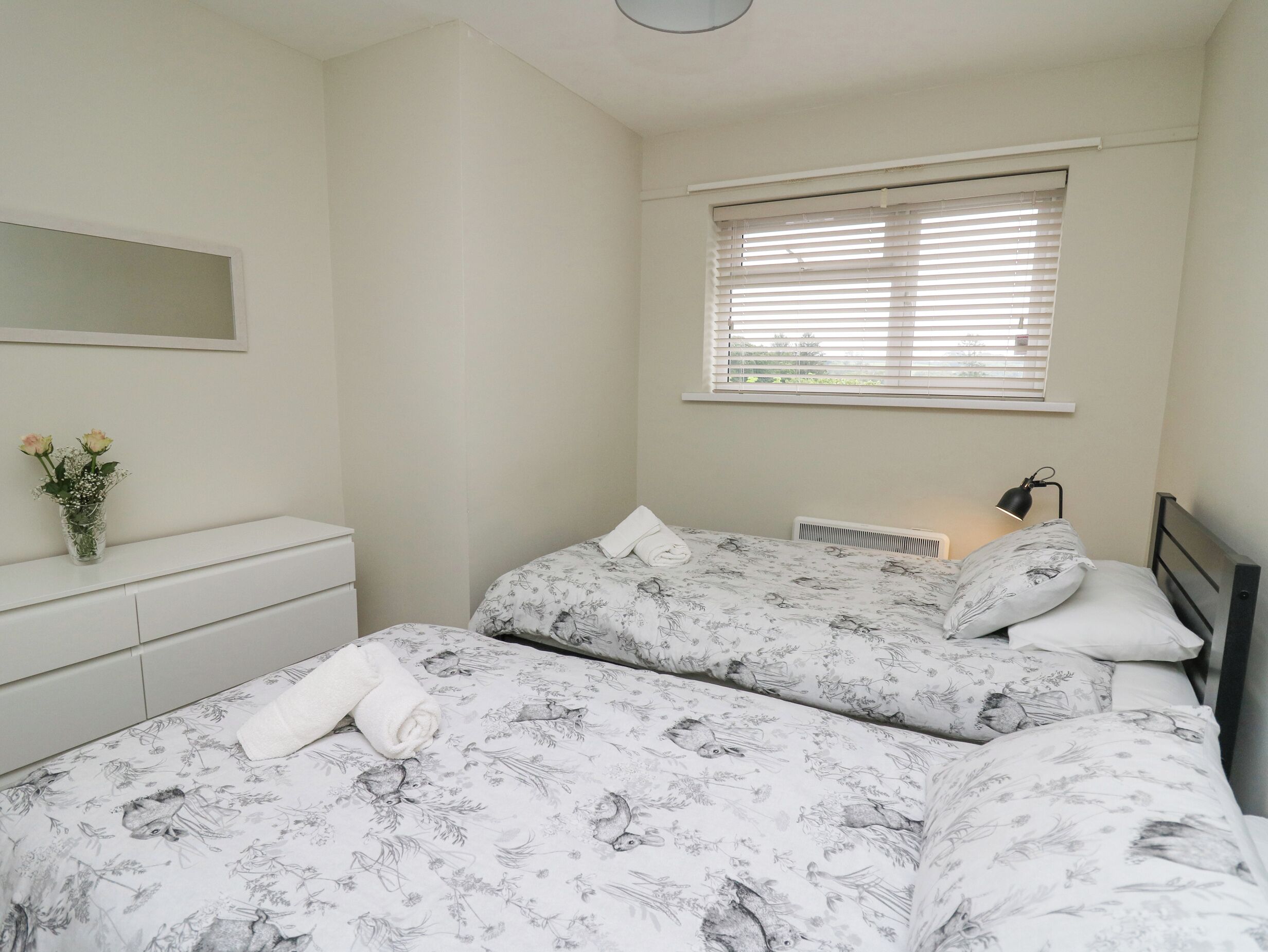 A bedroom with two beds, a bedside table, a mirror, and a window with blinds.