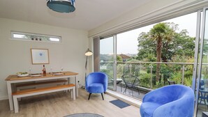 A modern living room with a dining area, large windows, and a balcony with a view of greenery.