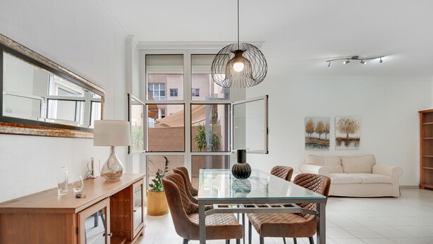 A modern living room with a glass-top dining table, brown upholstered chairs, a wooden cabinet, a hanging light fixture, and a large window with a view.