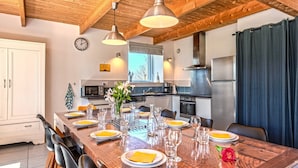 A dining area with a wooden table set for six, a kitchen in the background, and a clock on the wall. - Holiday Home with Private Heated Pool, Private Terrace, Garden and Wi-Fi (Plouguerneau)