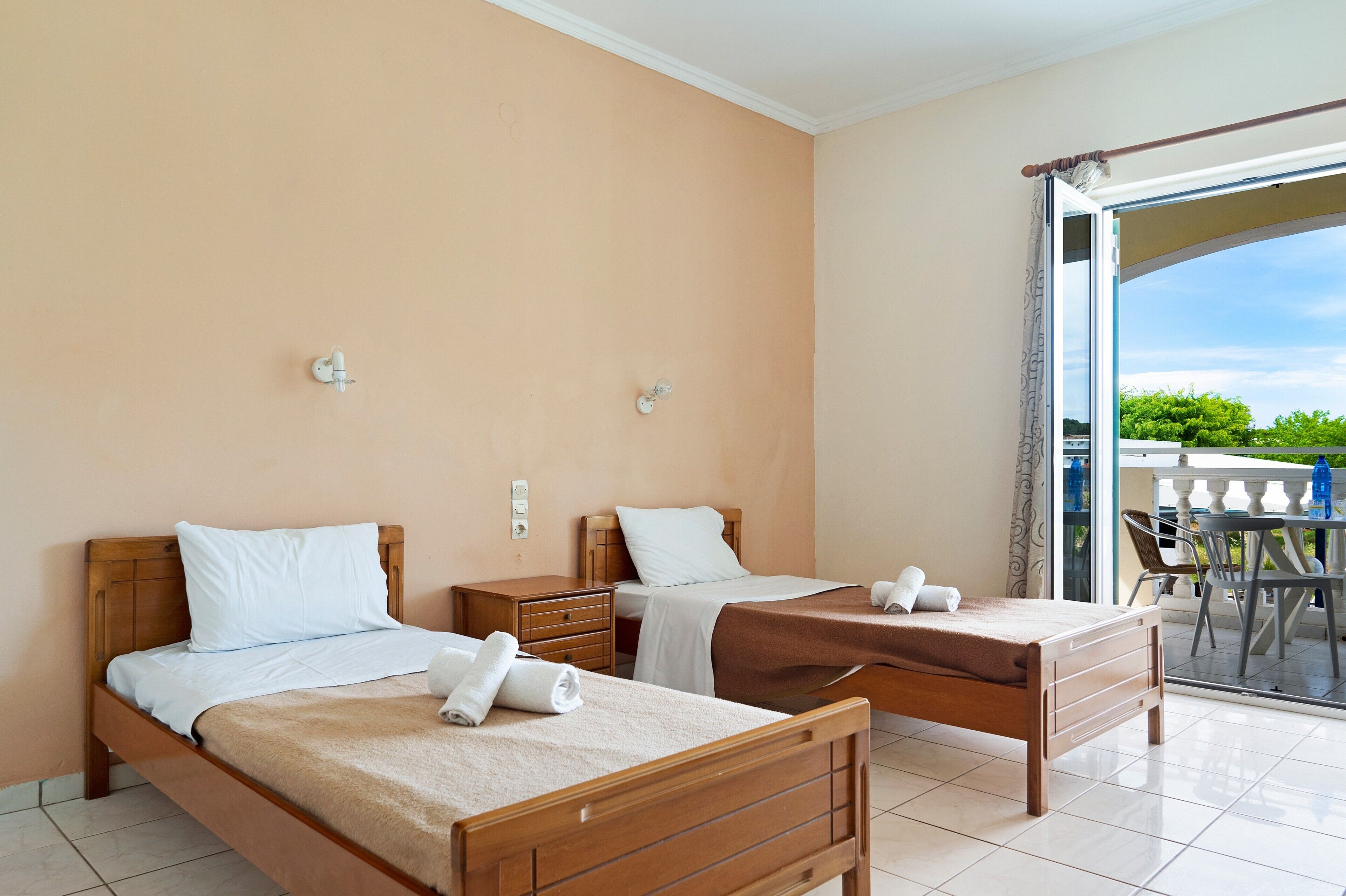 A hotel room with two wooden beds, a small table, and a balcony with chairs.
