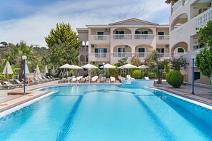 Una piscina con sillones de descanso y sombrillas, rodeada por un edificio de varios pisos con balcones.