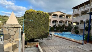 A stone structure, a neatly trimmed hedge, a swimming pool, and a multi-story building with balconies. - Studio 'Sandy Maria Studio 705' with Shared Pool, Wi-Fi (Zante)