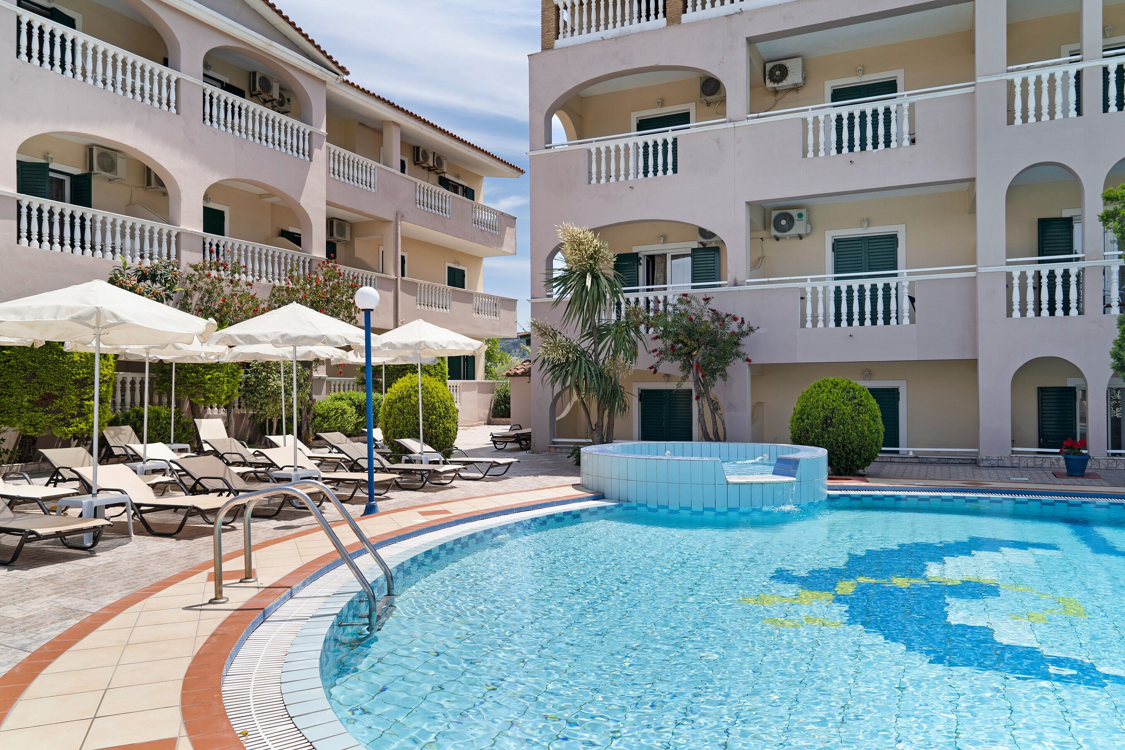 Una piscina amb gandules i ombrel·les, envoltada d'un edifici de diversos pisos amb balcons.