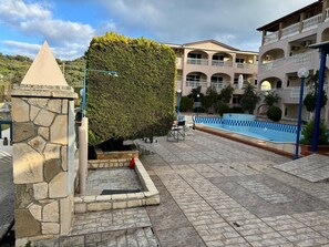 Una estructura de piedra, un seto bien cuidado, una piscina y un edificio de varios pisos con balcones.