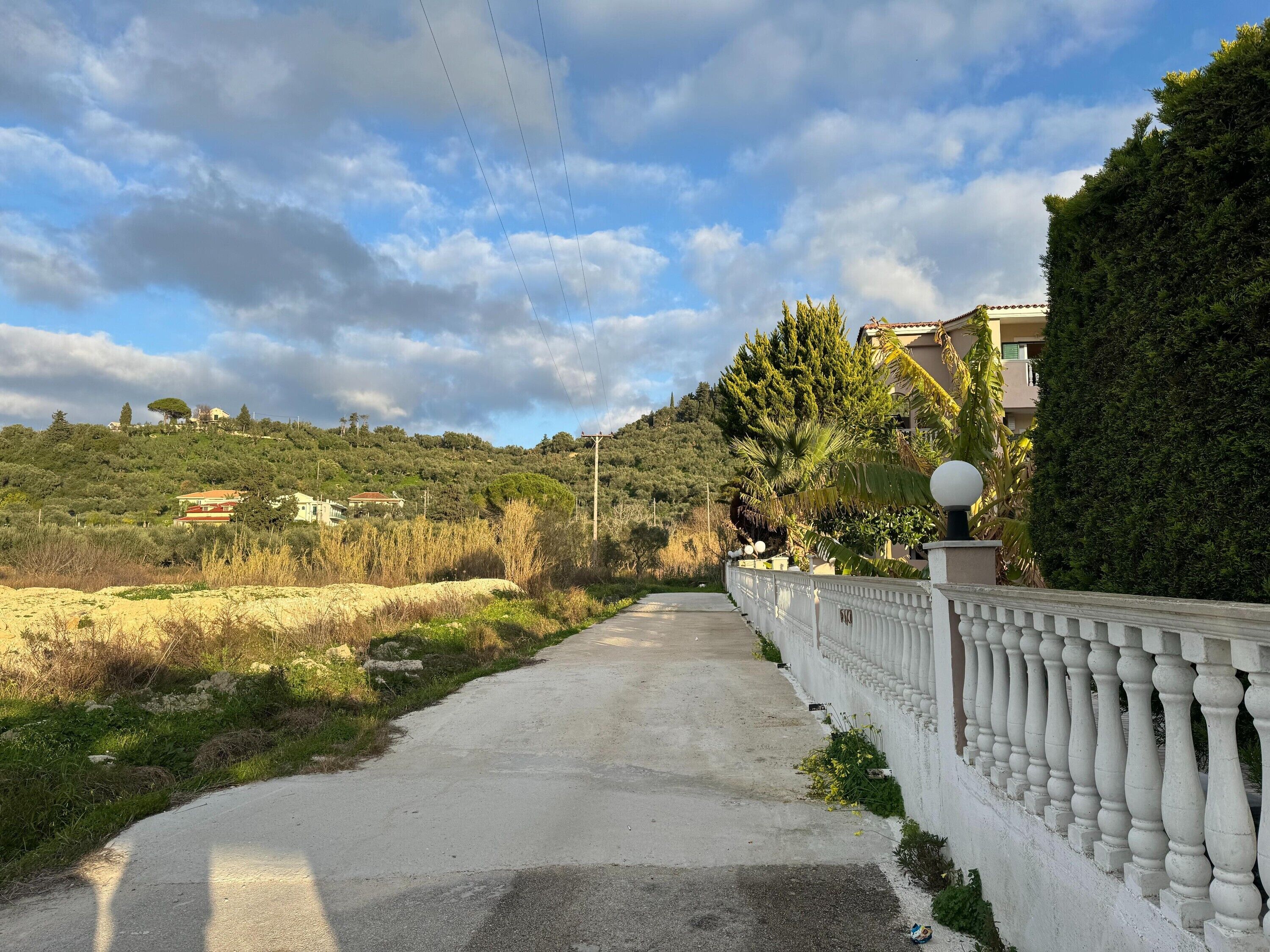 Un chemin pavé, bordé d’une balustrade blanche, mène à un quartier résidentiel où se trouvent des maisons et une végétation luxuriante.