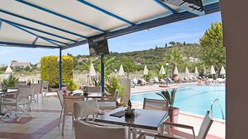 Un espace extérieur aménagé, abrité et doté de tables et de chaises, une piscine et une vue sur une colline.
