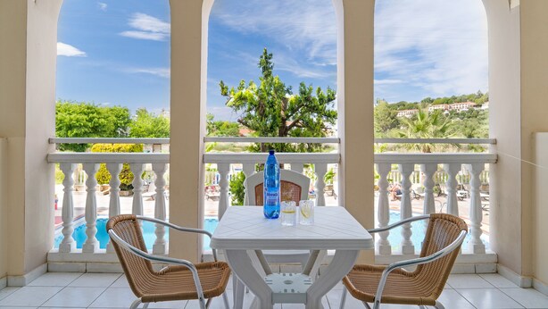 Un balcon avec vue sur la piscine, une table blanche, des chaises en osier et une bouteille d’eau bleue.