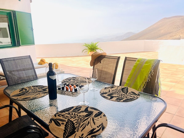 A glass-top table with chairs, a bottle, and glasses on a terrace with a view of mountains.