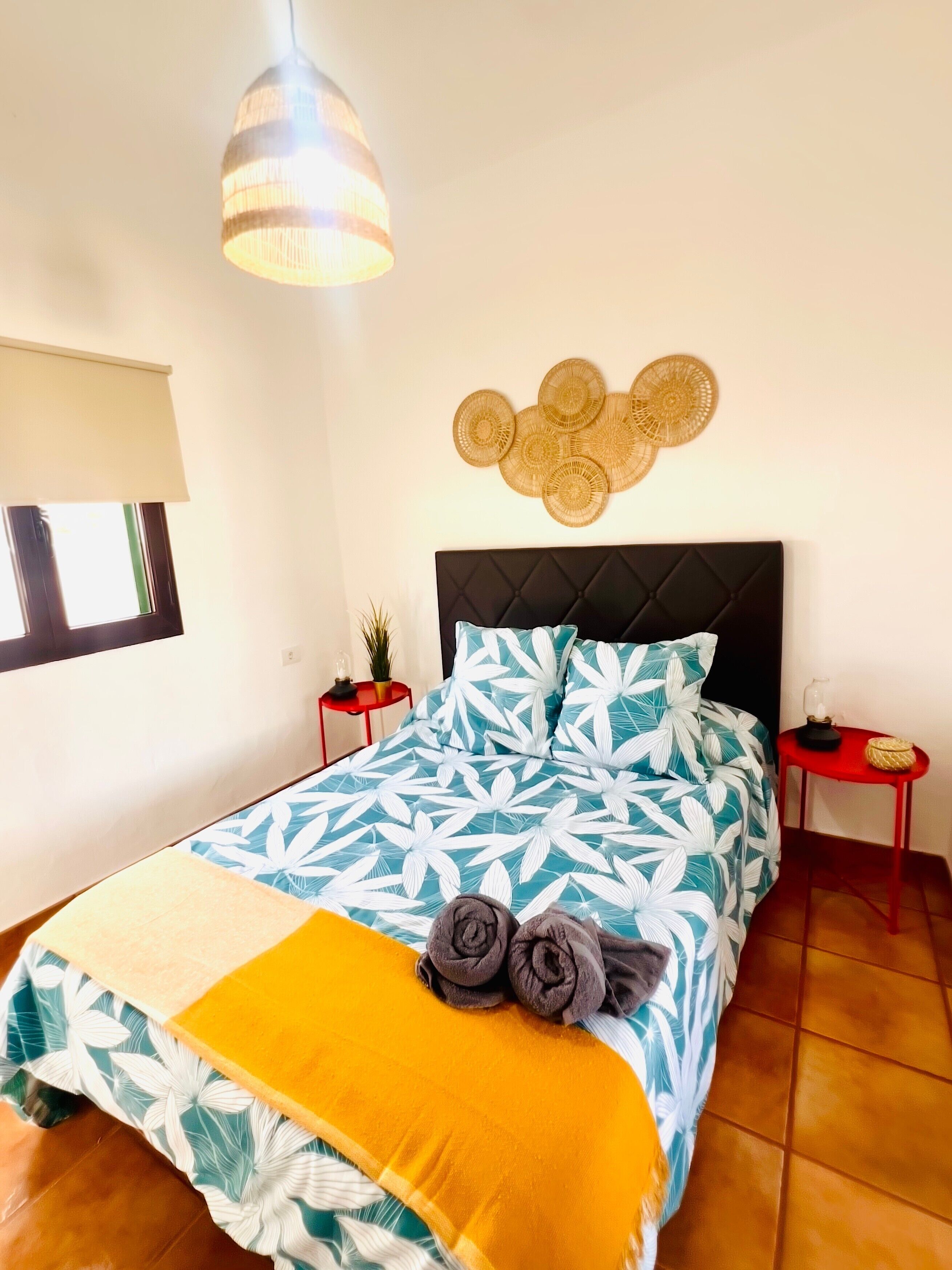 Un dormitorio con cama, una lámpara colgante, decoración en la pared, una mesita y una ventana.