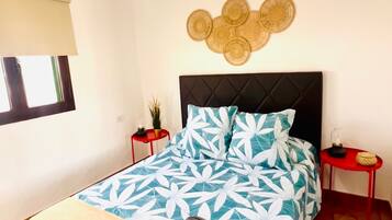 Un dormitorio con cama, una lámpara colgante, decoración en la pared, una mesita y una ventana.