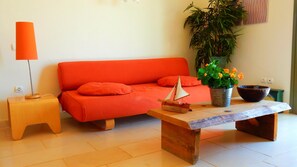 A bright orange sofa with matching cushions, a small wooden table with a model sailboat, a potted plant with orange flowers, and a metal bucket.