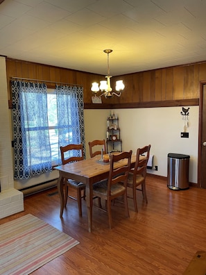 Un coin repas avec une table et des chaises en bois, un lustre, une fenêtre ornée de rideaux à motifs et une étagère murale sur laquelle sont disposés divers objets.