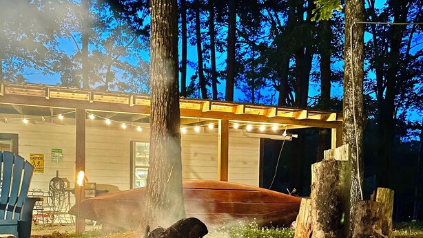 Un espace extérieur rustique aménagé avec des sièges, un foyer entouré de rochers et de troncs d’arbres, des bancs en bois et une pergola en bois décorée de guirlandes lumineuses.