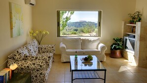 Una sala de estar con un sofá, una mesa de centro con una botella y vasos, un televisor y una ventana con vistas a una colina.