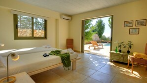 Un dormitorio con cama, lámpara de noche y un banco. Se ve una ventana con vistas a árboles y un muro de piedra.