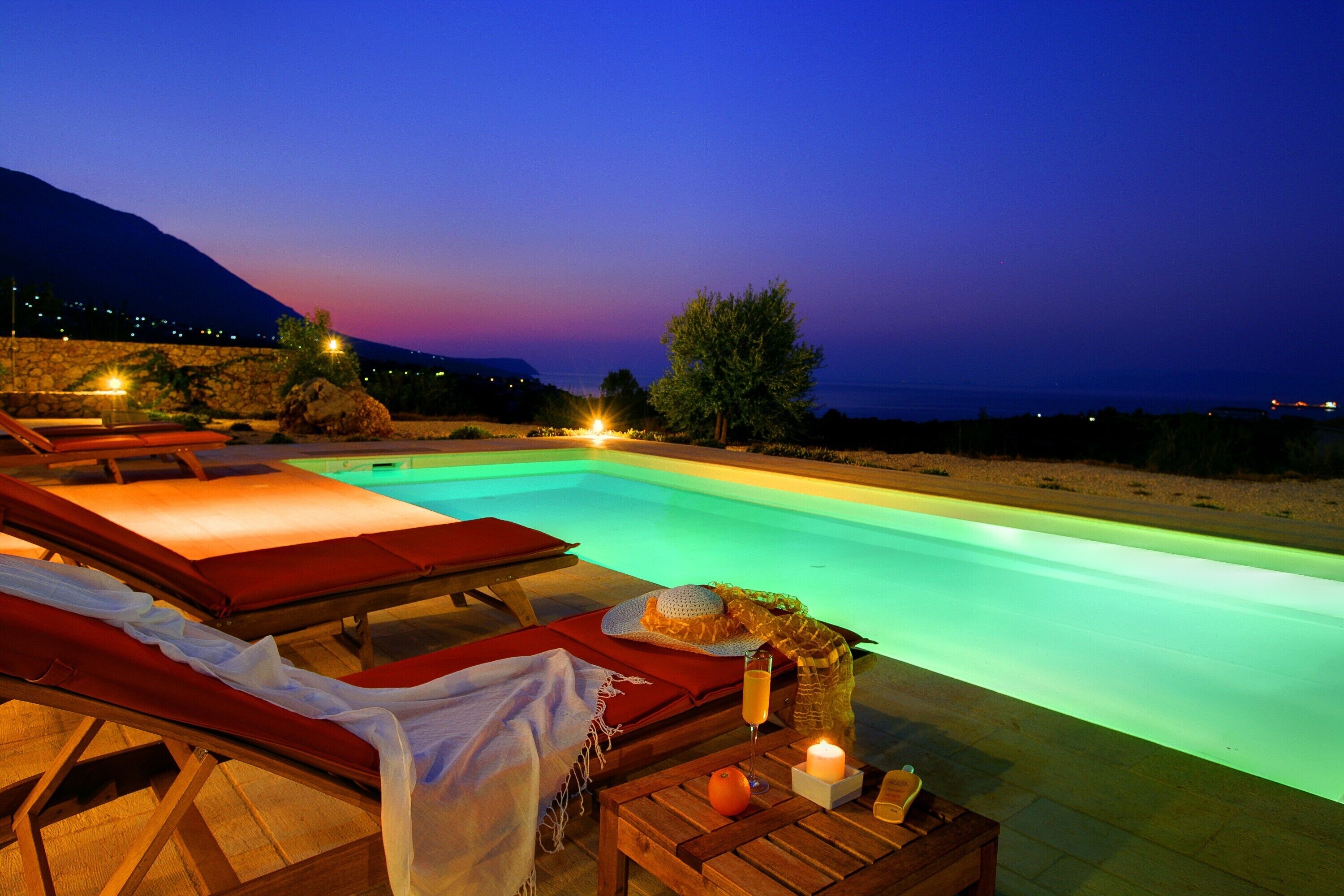 Un área junto a la piscina con sillones, una mesa con vela y vista al mar al anochecer.