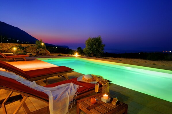 A poolside area with lounge chairs, a table with a candle, and a view of the sea at dusk.