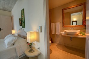 Et soveværelse med seng, sengeborde, væglampe, et billede på væggen, badeværelse med vask, spejl, håndklædetørrestativ og toilet.