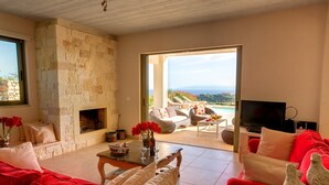 A living room with a stone fireplace, red sofas, a television, and a view of a pool and mountains. - Villa 'Corali Ideales Resort' with Sea View, Wi-Fi and Air Conditioning (kefalonia)