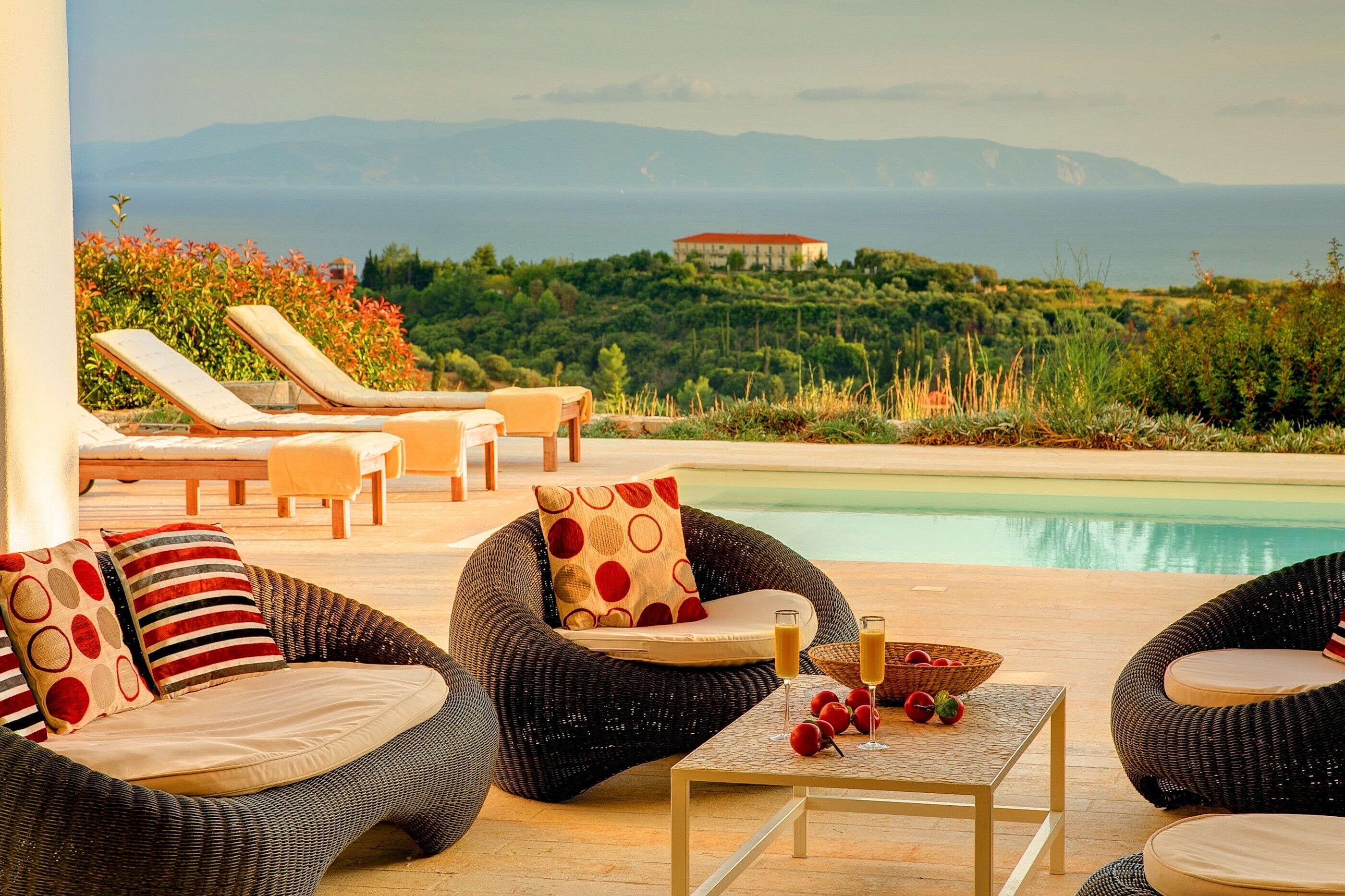 Zona piscina con mobili in vimini, un tavolo con frutta e bevande, e vista sul mare e sulle colline.