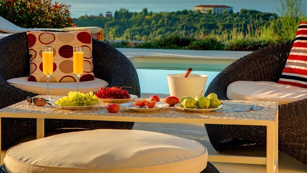 A wicker lounge chair with a cushion, a table with fruit and drinks, and a view of a pool and greenery. - Villa 'Corali Ideales Resort' with Sea View, Wi-Fi and Air Conditioning (kefalonia)