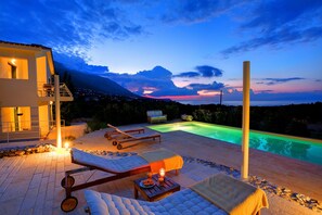 Ett modernt utomhusområde med en pool, solstolar, ett bord med ljus och utsikt över bergen i skymningen.