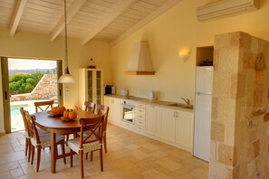 A kitchen with a dining table, chairs, a hanging light fixture, a stone wall, and a view of greenery outside.