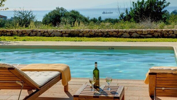 Zona junto a la piscina con sillas de descanso, una mesa con una botella y vasos, y un muro de piedra.