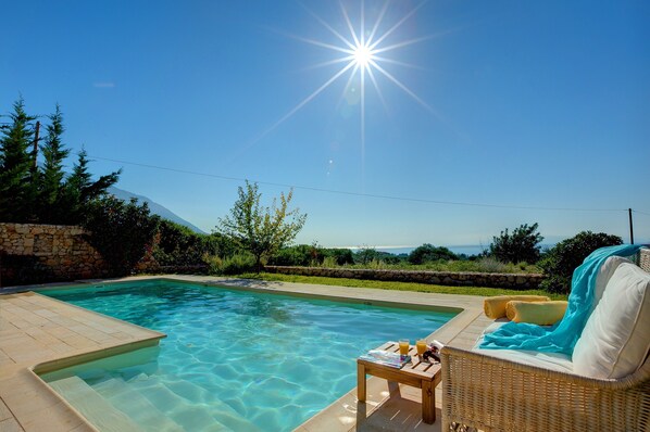 Une piscine baignée de soleil, un mur de pierre et un fauteuil de jardin en osier avec des serviettes.