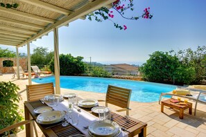 Un área de comedor junto a la piscina con una mesa dispuesta para dos, sillas y vistas a la piscina y al paisaje.